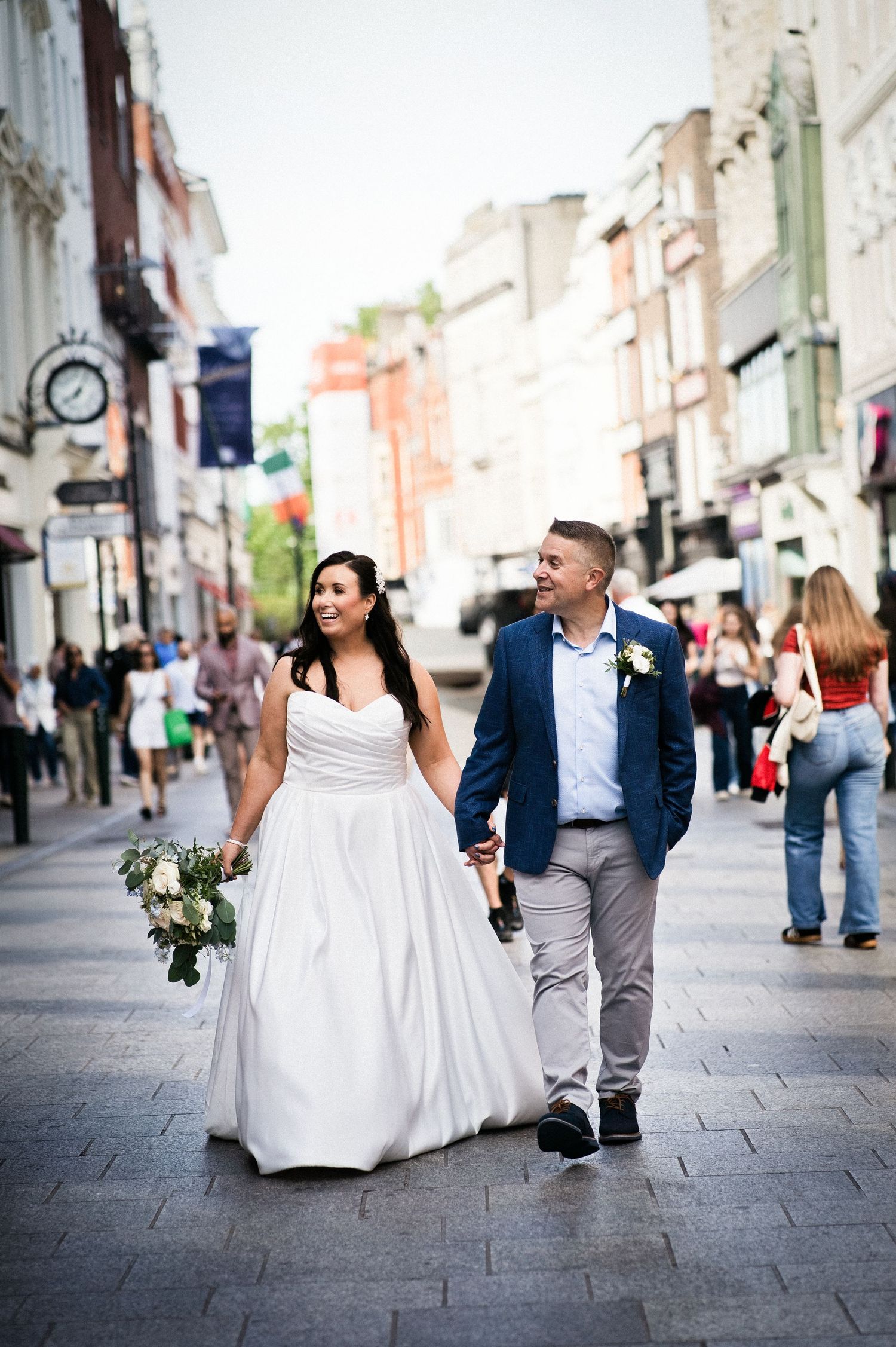 Bride and groom walking hand-in-hand through a bustling city street, bride holding a white bouquet in a flowing gown.
