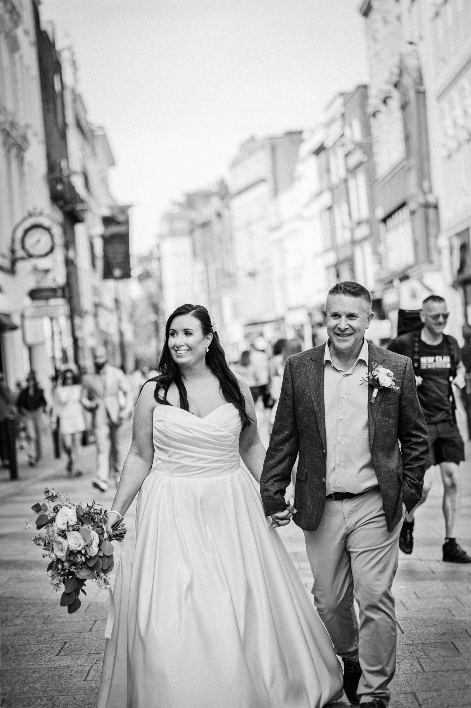 A smiling bride and groom walk hand-in-hand through a busy city street, she holding a bouquet in black and white.