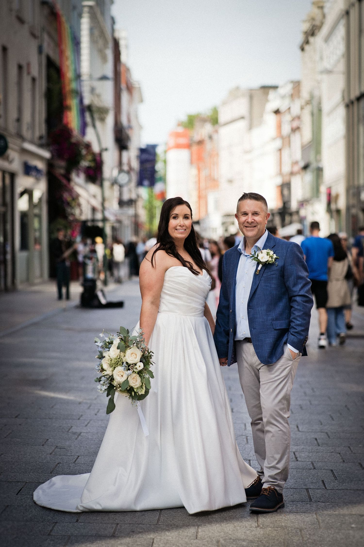 Bride and groom pose on a bustling city street, bride holding white bouquet in elegant gown, groom in blue blazer.