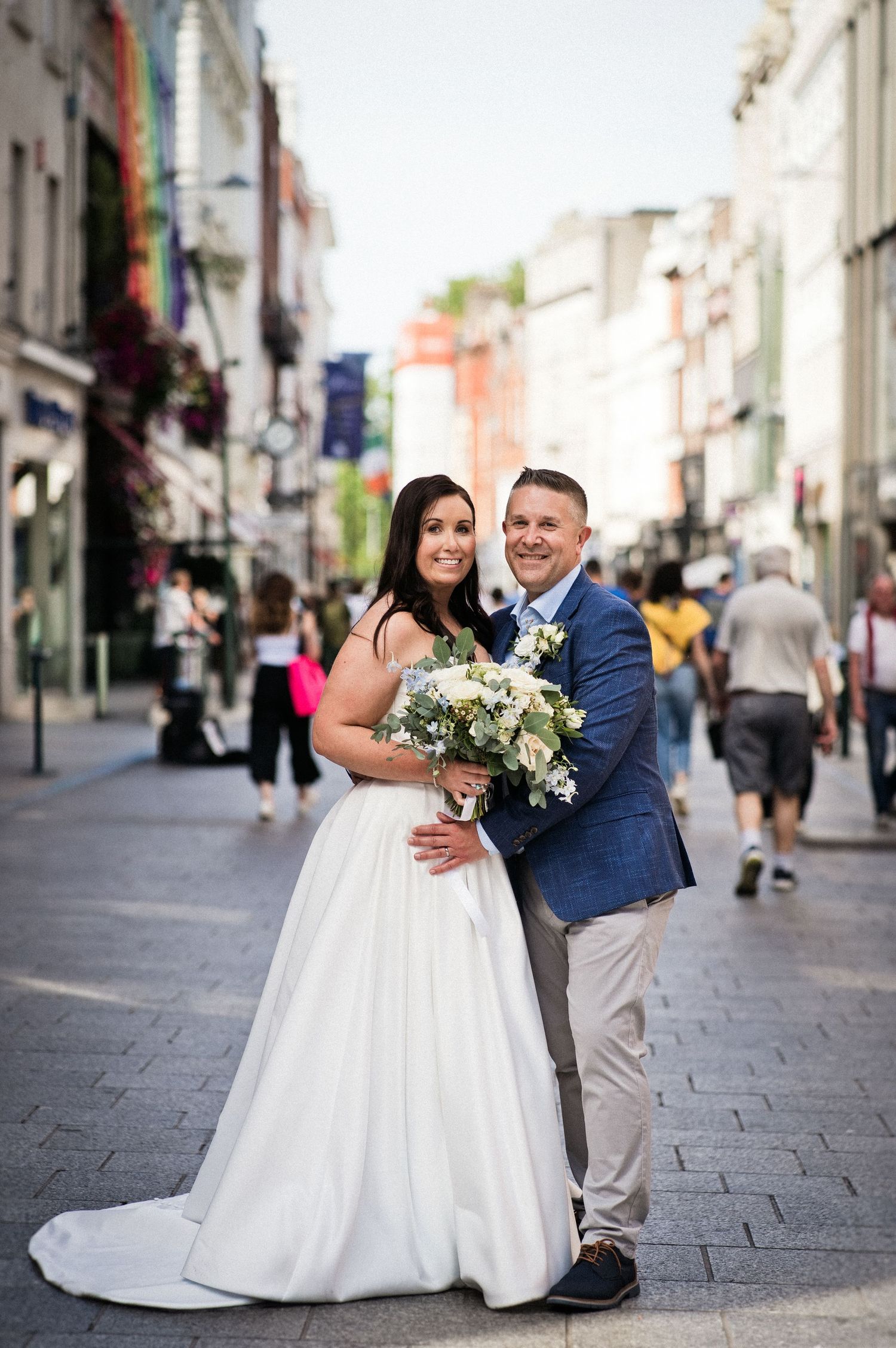 Bride and groom pose on a cobblestone city street, bride holding white bouquet in elegant gown.