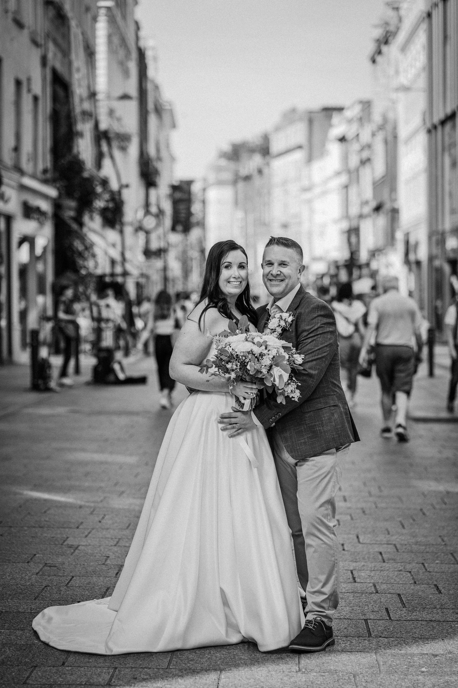 Black and white photo of a bride and groom posing on a busy city street, bride holding a bouquet.