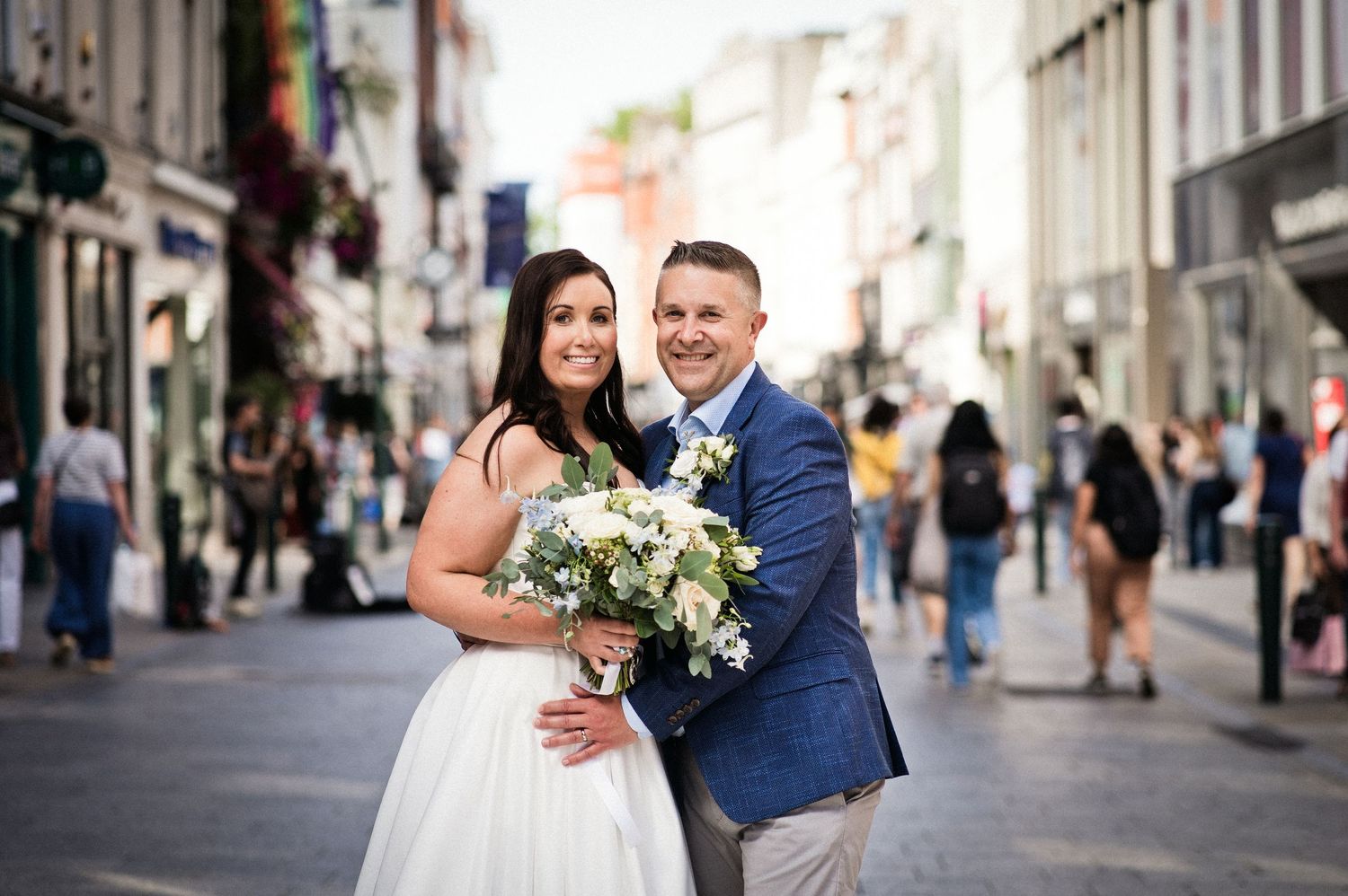 Bride and groom pose together on a busy pedestrian street, bride holding white bouquet, groom in blue blazer.