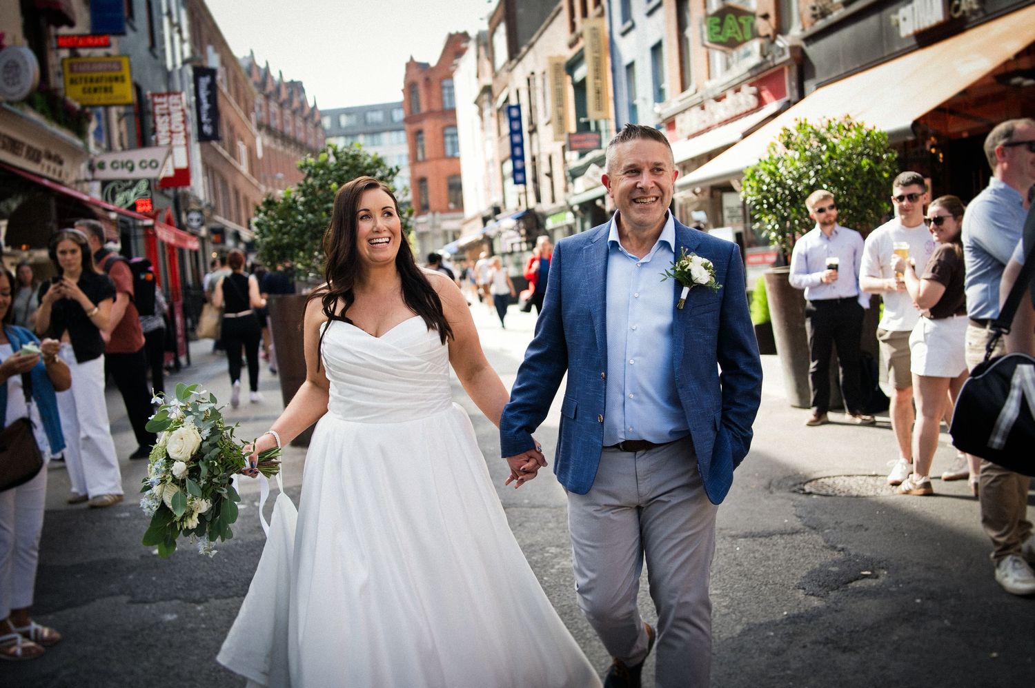 Newlywed couple walking hand-in-hand on a busy city street, bride in white gown holding bouquet, groom in blue blazer.