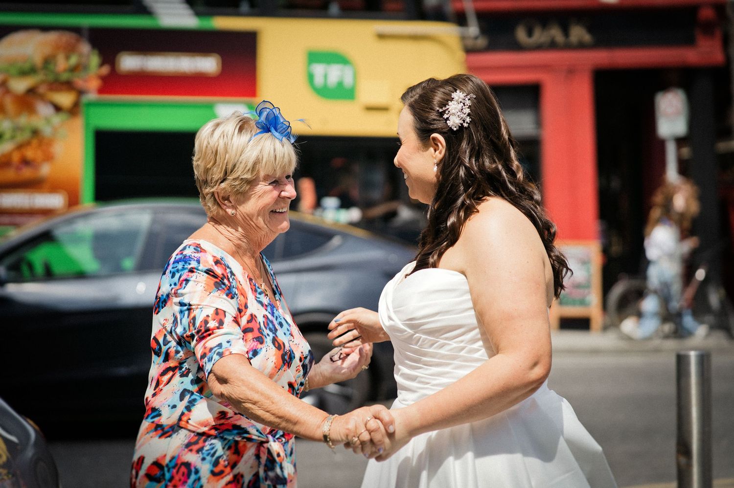 A bride in a white gown shares a joyful moment with an older woman on a busy street.