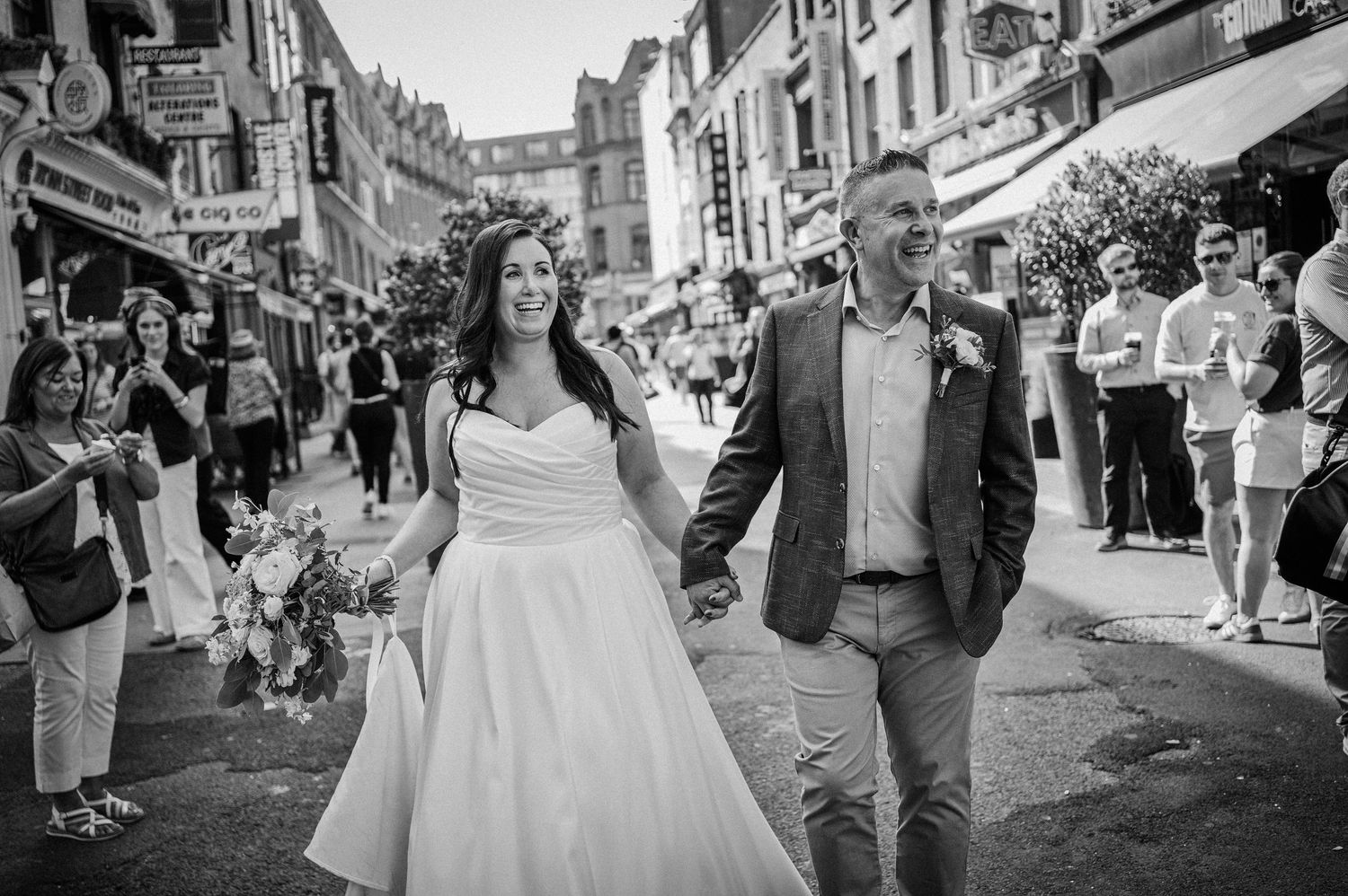 Black and white photo of a bride and groom holding hands walking through a busy city street on their wedding day.