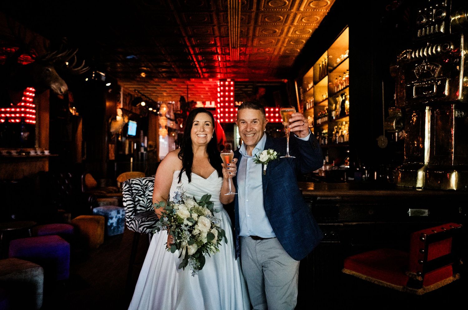 Bride and groom toasting with champagne glasses in a dimly lit bar venue on their wedding day.