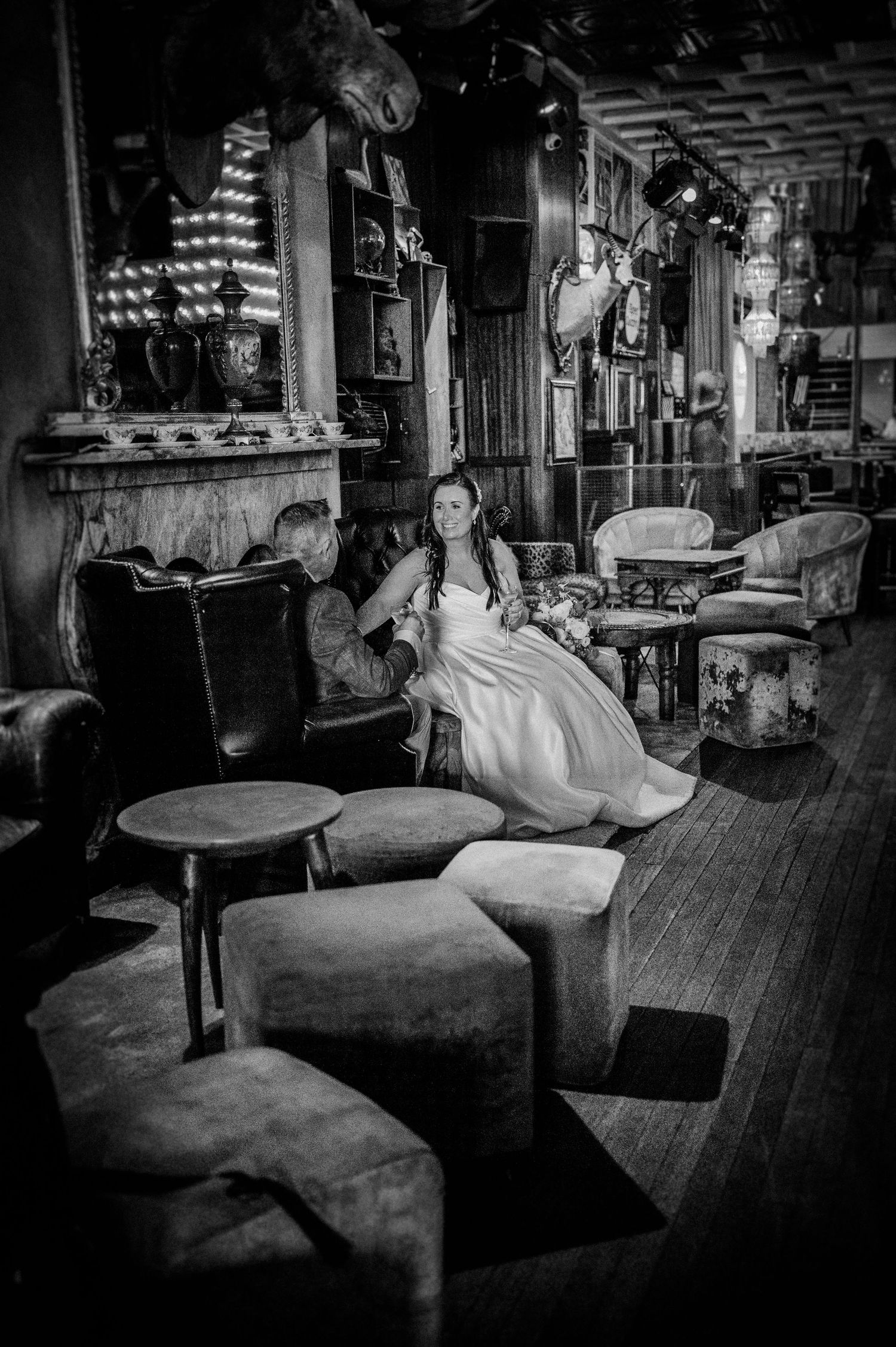 Black and white photo of a bride in a white dress sitting in an ornate lounge chair inside a rustic, dimly lit venue.