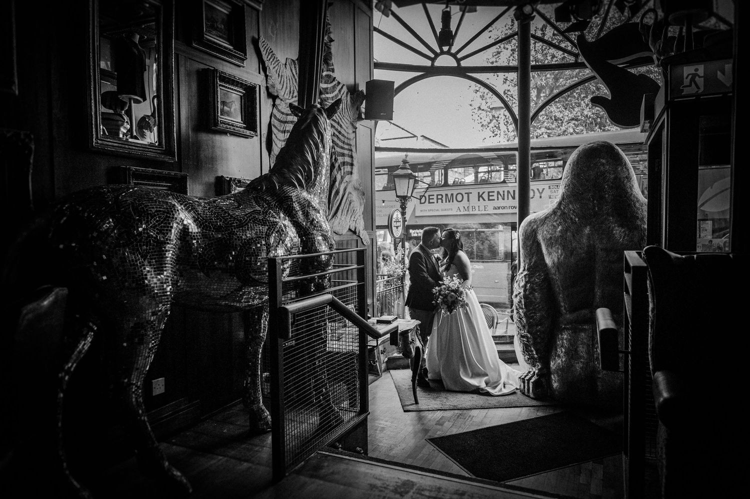 Black and white wedding photo with bride and groom framed by decorative horse sculptures in a vintage venue.