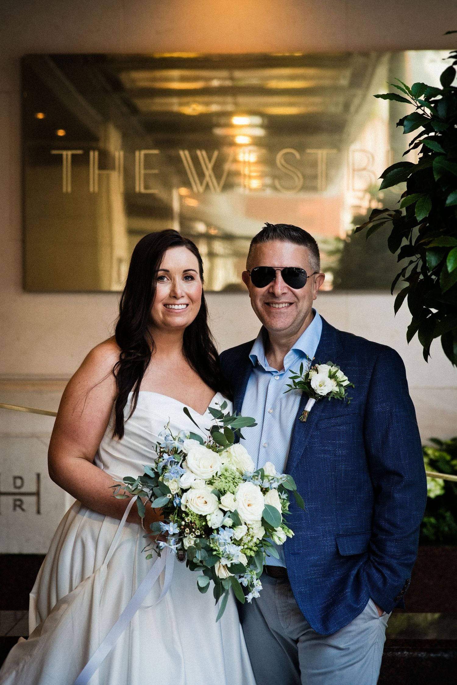 Bride in white gown holding white floral bouquet poses with groom in blue suit outside The Westin hotel.