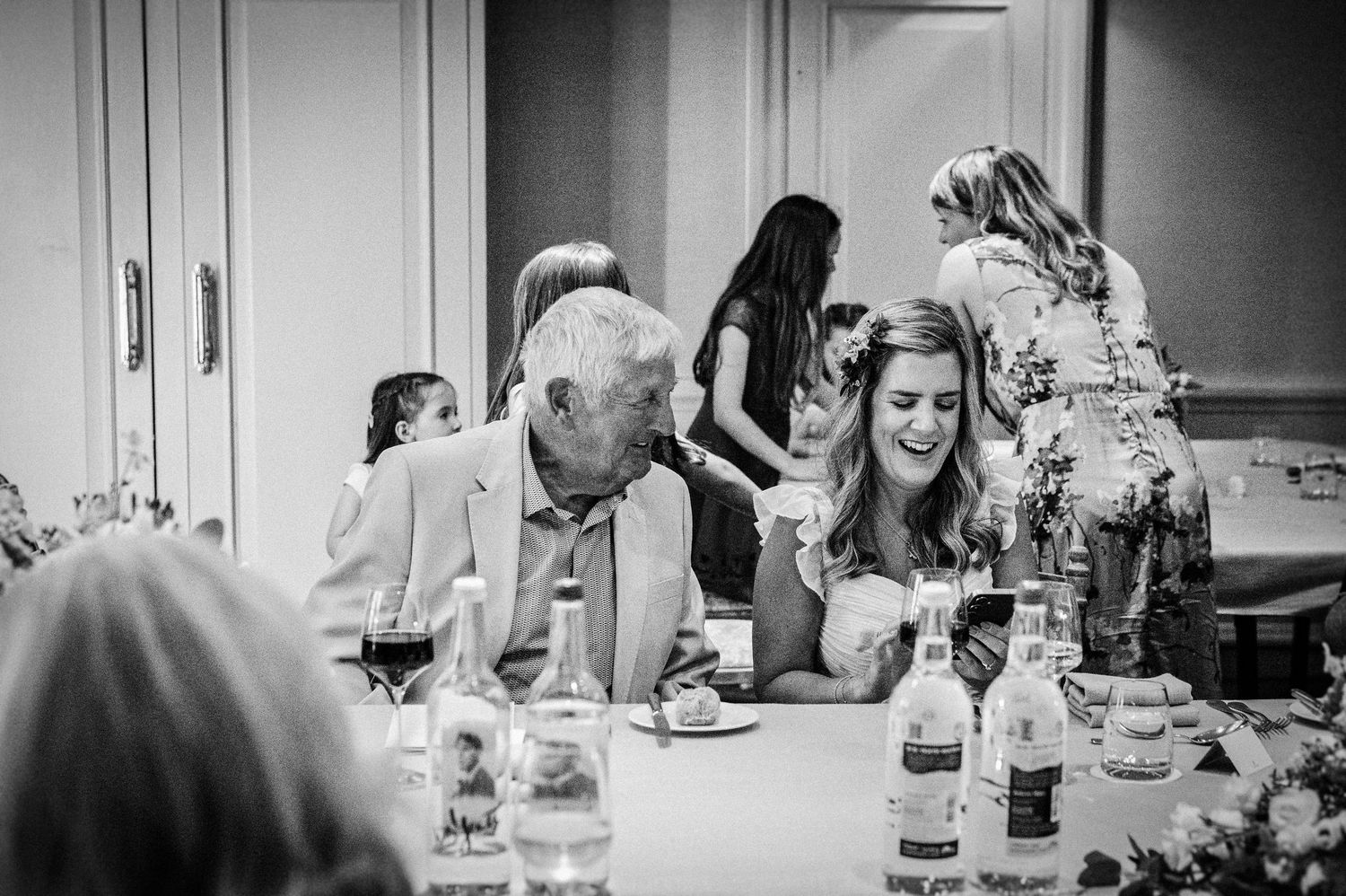 Black and white photo of guests laughing and chatting at a wedding reception dinner table.