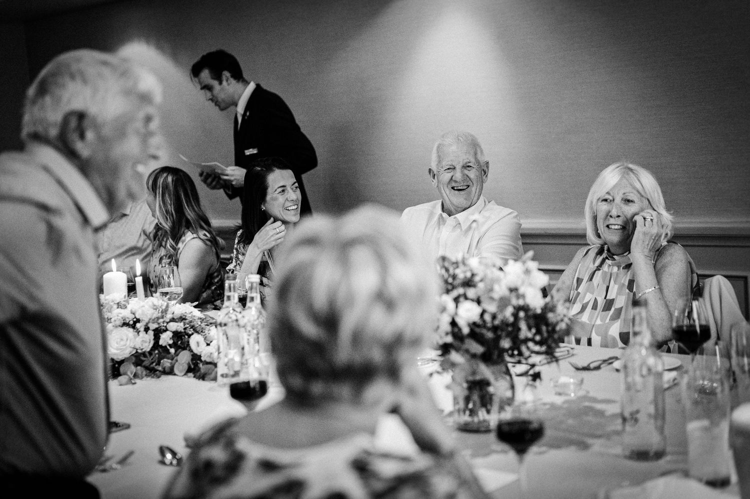 Black and white wedding reception photo showing guests laughing and enjoying dinner at elegantly decorated tables with floral centerpieces.