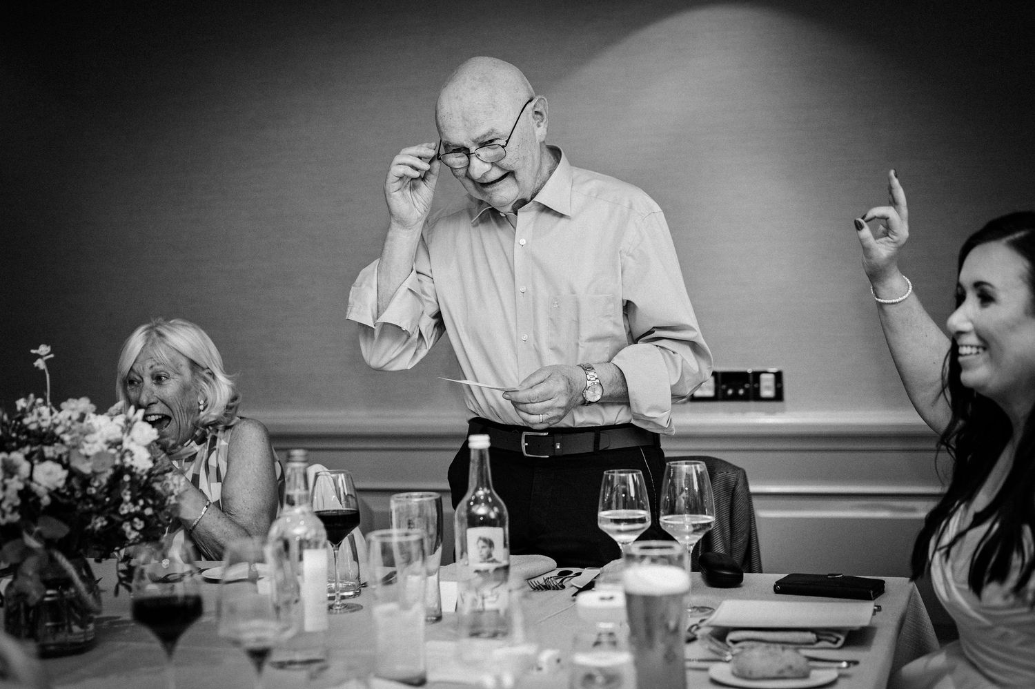 Elderly man adjusts glasses at formal dinner table while guests laugh in black and white wedding reception photo.