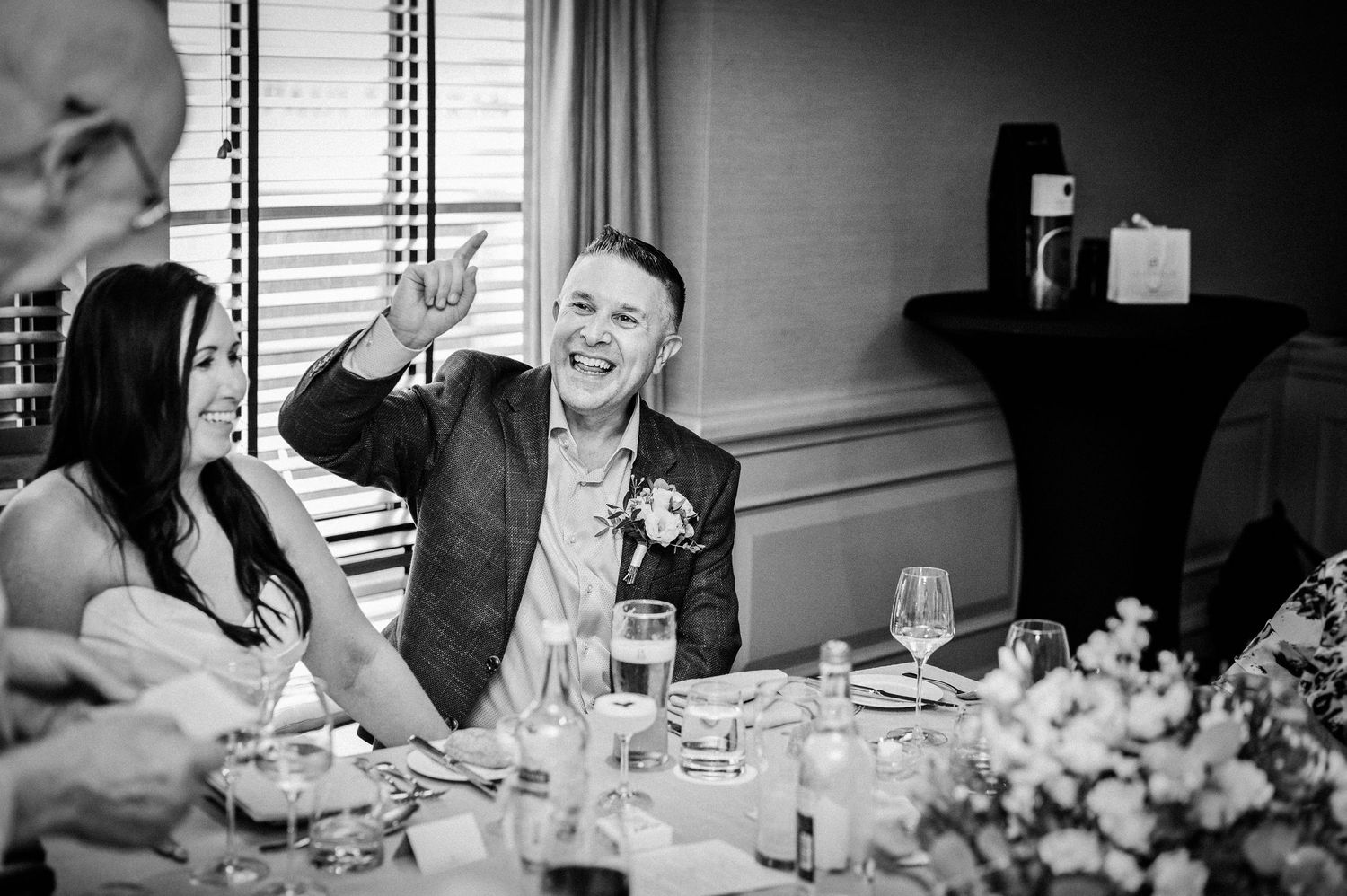 Man laughing joyfully at wedding reception table, raising hand while seated next to bride in black and white photo.