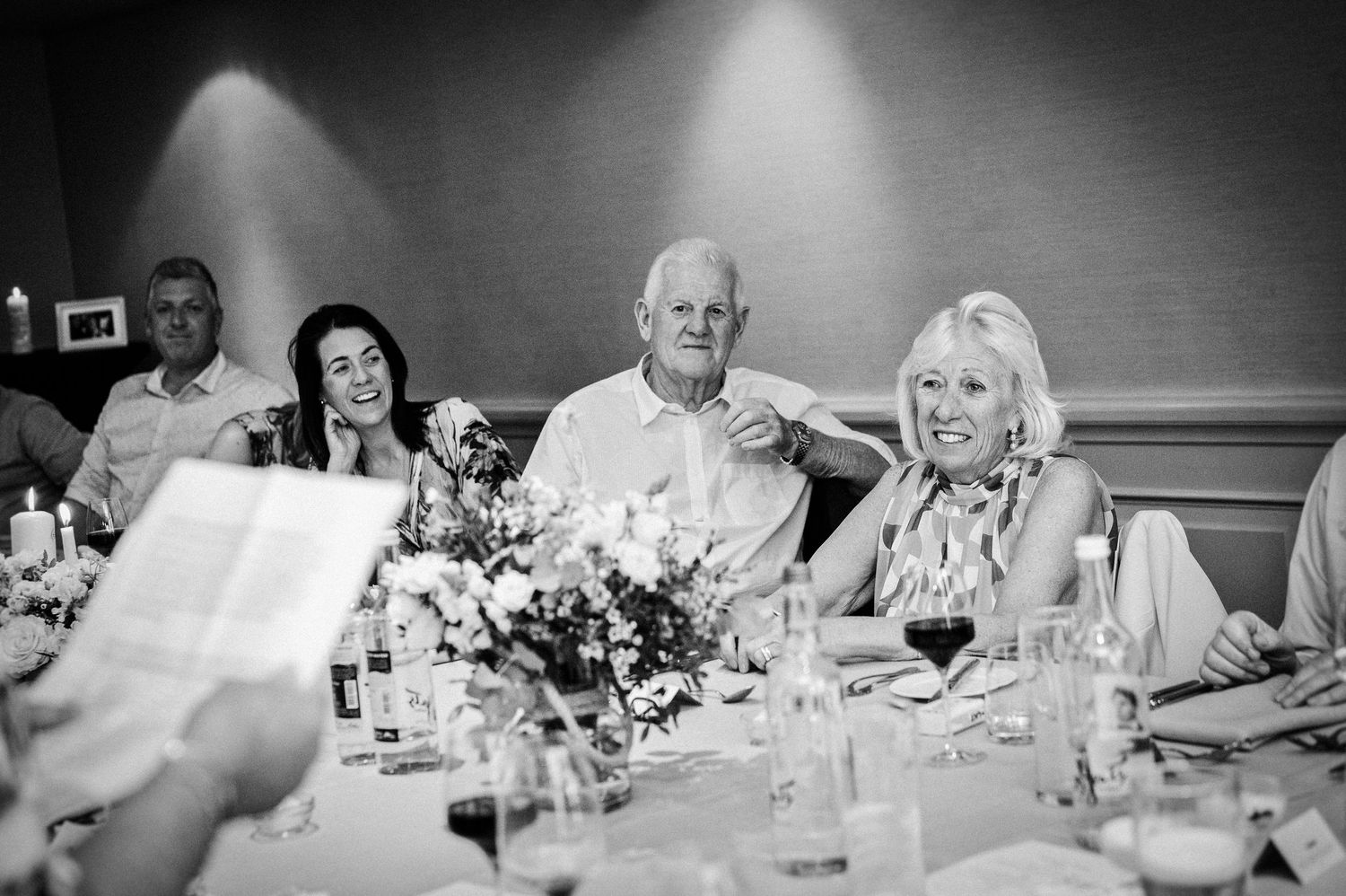Guests laughing and smiling at a wedding reception dinner table with floral centerpieces in a black and white photo.