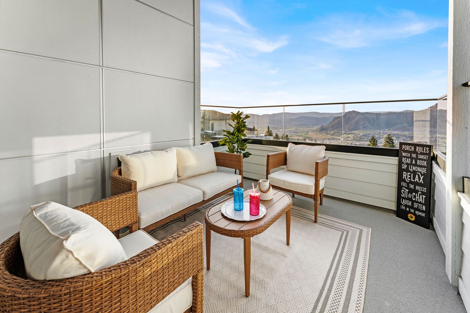 Modern balcony with wicker furniture, white cushions, coffee table, and panoramic mountain views under a clear blue sky.