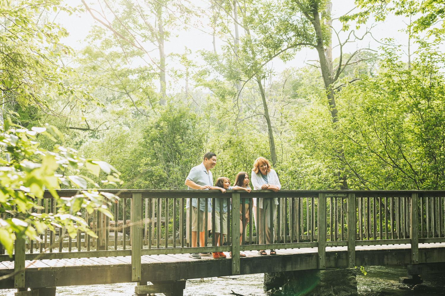Two people enjoying a sunny day on a wooden bridge surrounded by lush green trees over a peaceful stream.