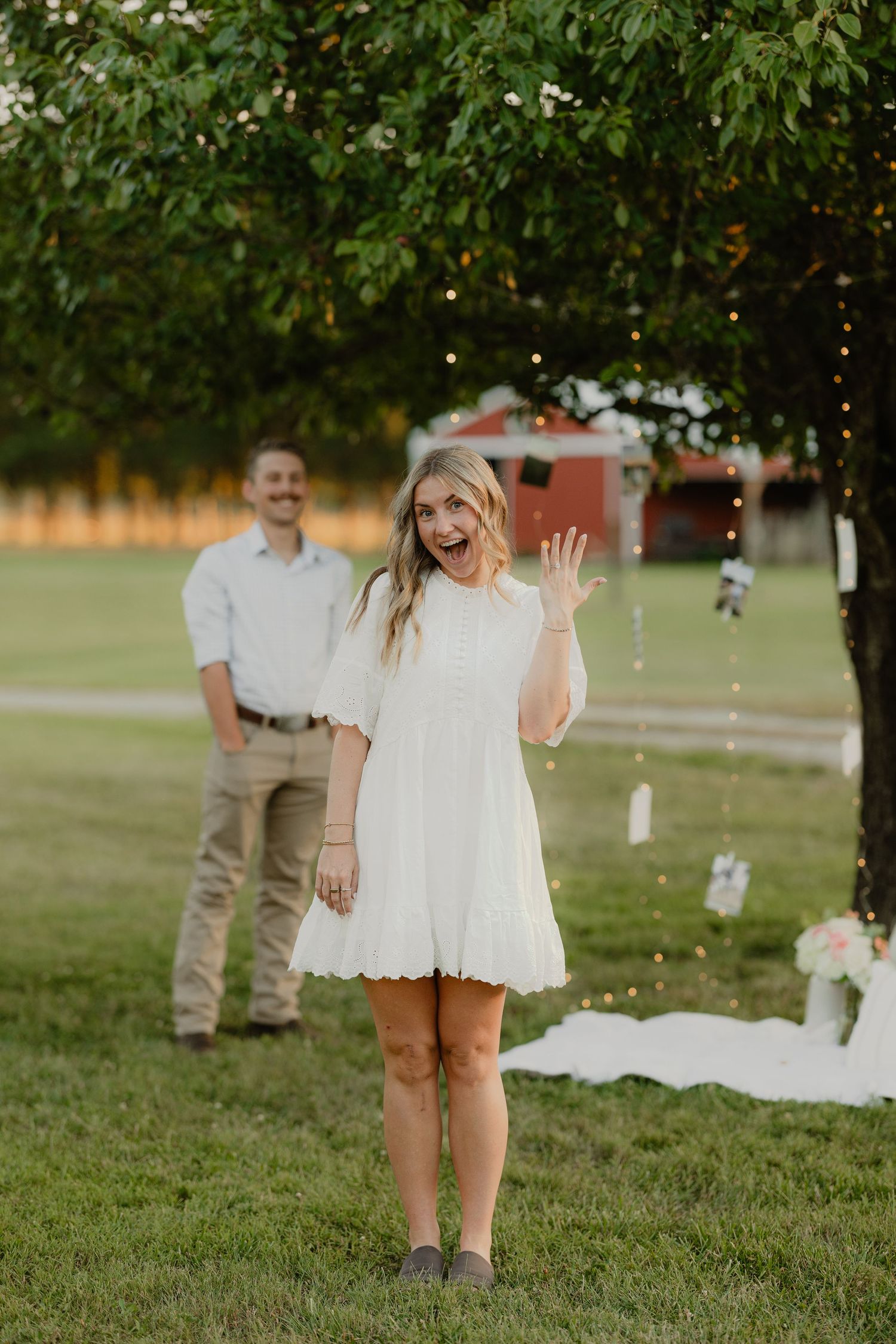Excited bride in white dress shows off engagement ring outdoors with groom smiling behind her.