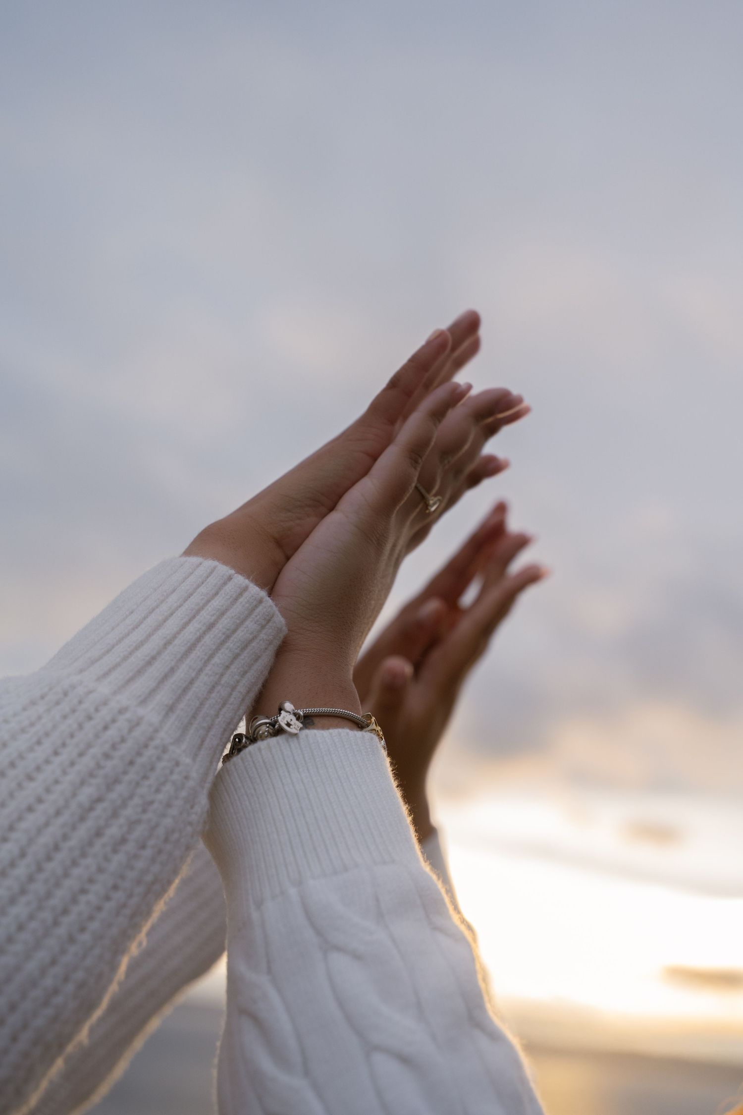 Two hands raised toward a cloudy sunset sky, wearing a silver charm bracelet and white knit sweater sleeves.