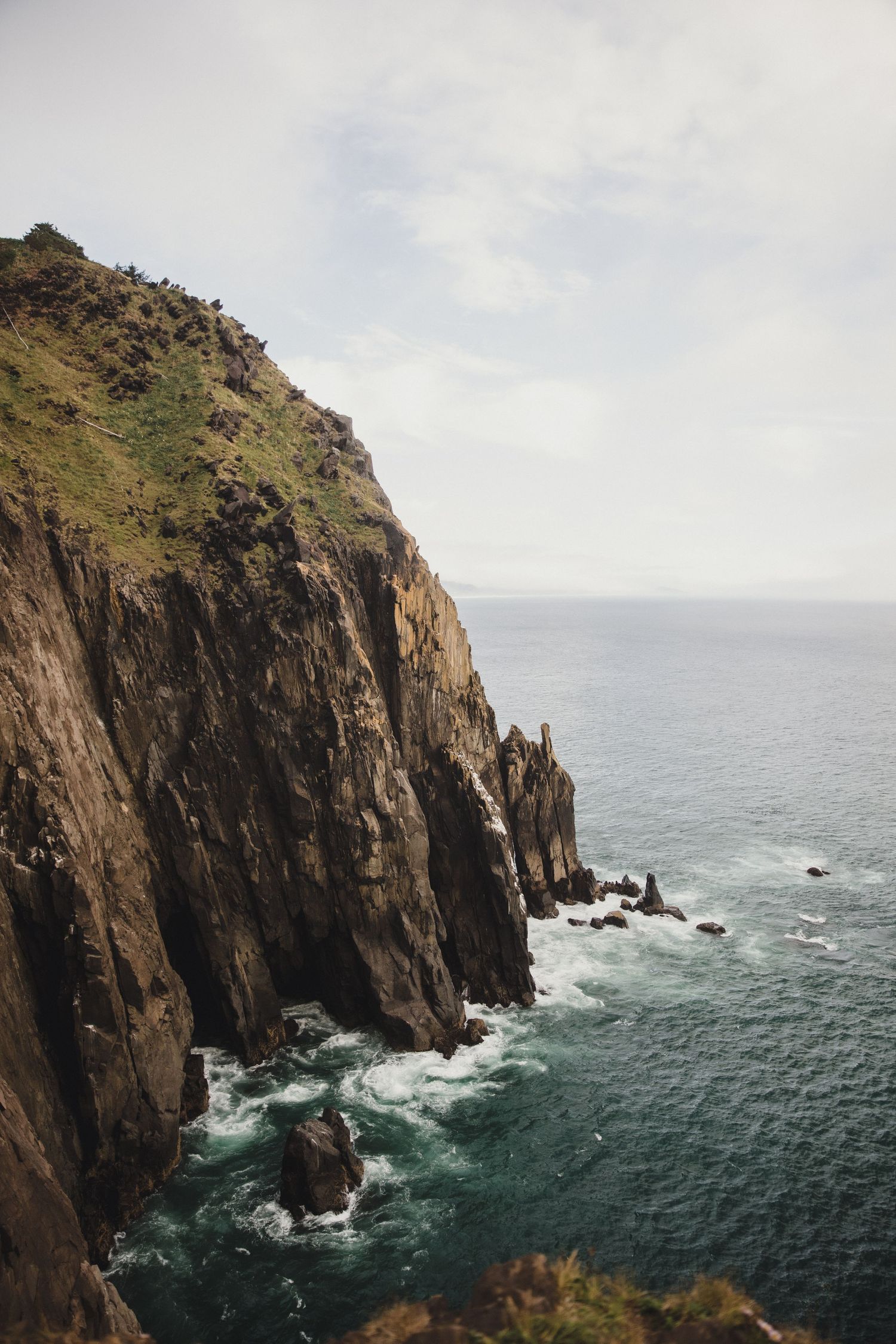 Dramatic coastal cliffs with green summit drop sharply to churning turquoise ocean waves below under overcast sky.