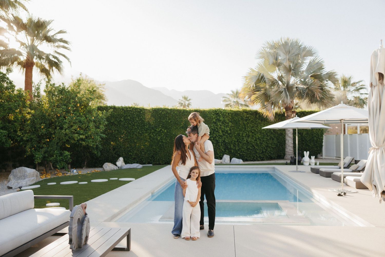 Family of three embracing by a luxurious outdoor pool with palm trees and mountain views in Palm Springs.