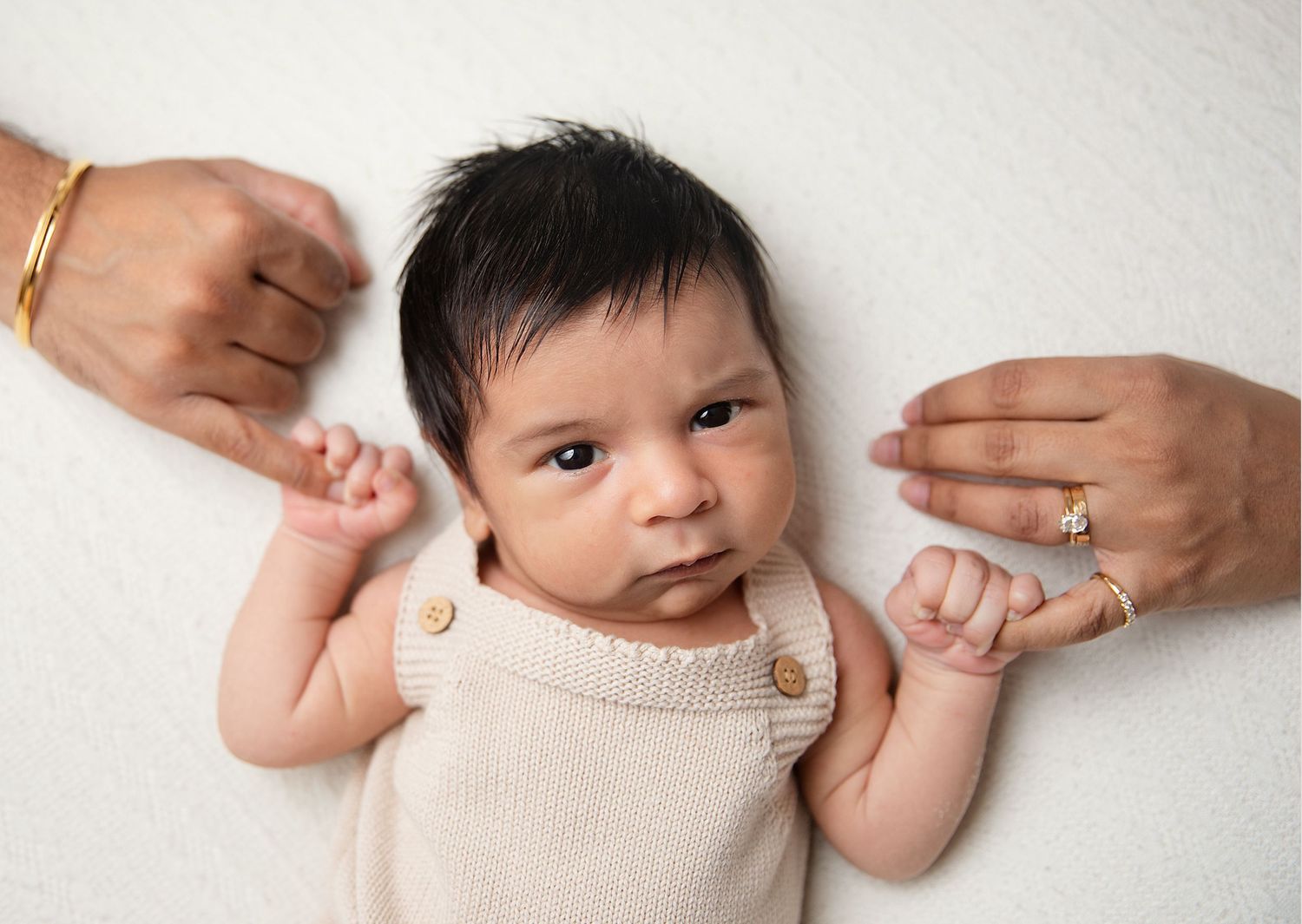 Newborn baby in beige romper lying on white background, held by two adults, during a newborn session in South East London