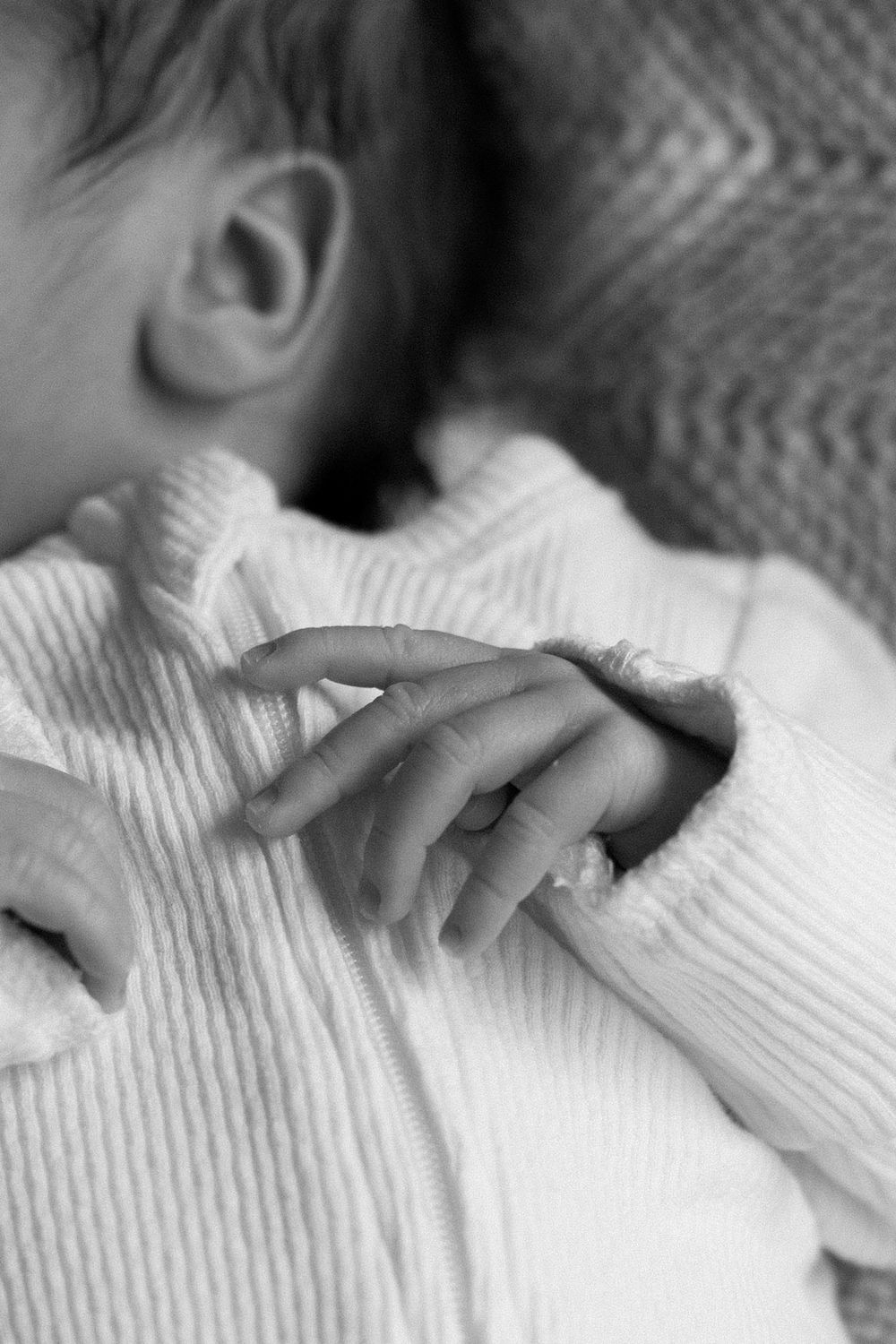 Black and white close-up of a sleeping newborn baby's tiny hand resting on a white ribbed onesie.