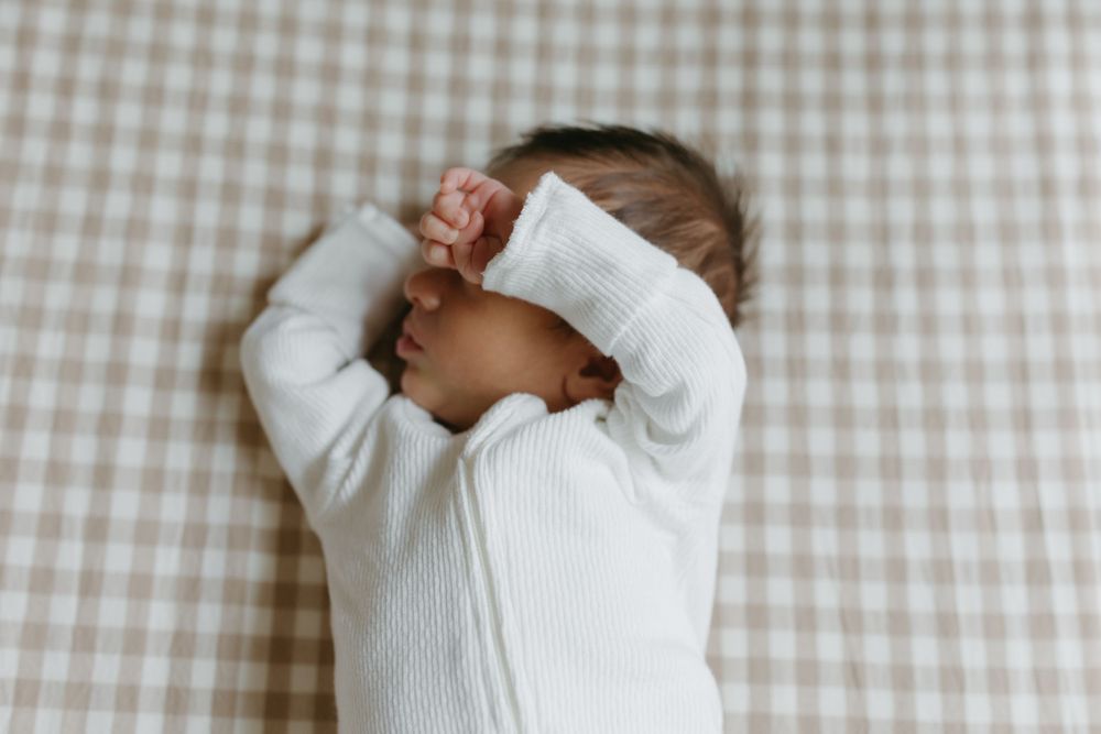 Newborn baby sleeping peacefully in white zip-up onesie on beige gingham blanket with arms raised.