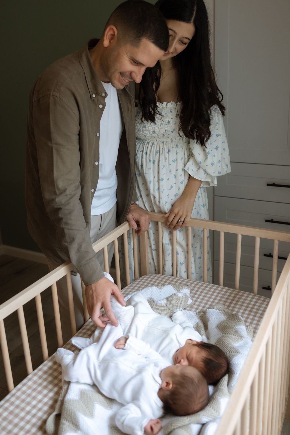 New parents smile lovingly at their newborn twins lying in a wooden crib.