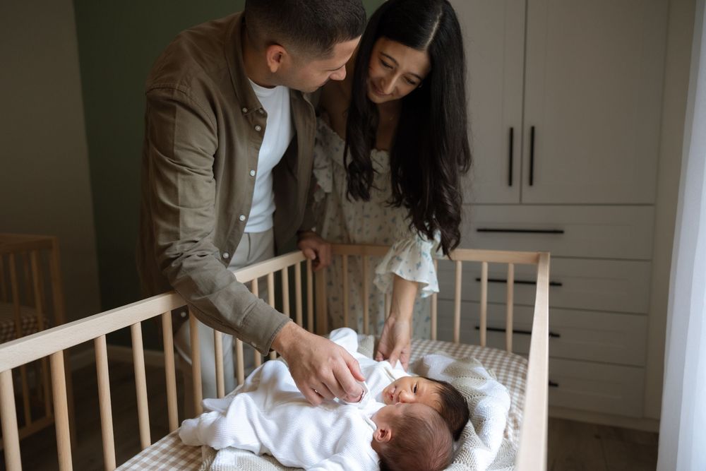Parents lovingly tend to their newborn baby lying in a wooden crib in a softly lit nursery room.