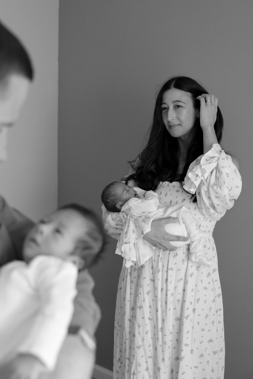 Mother in floral dress holds newborn while father cradles another baby in black and white photo.