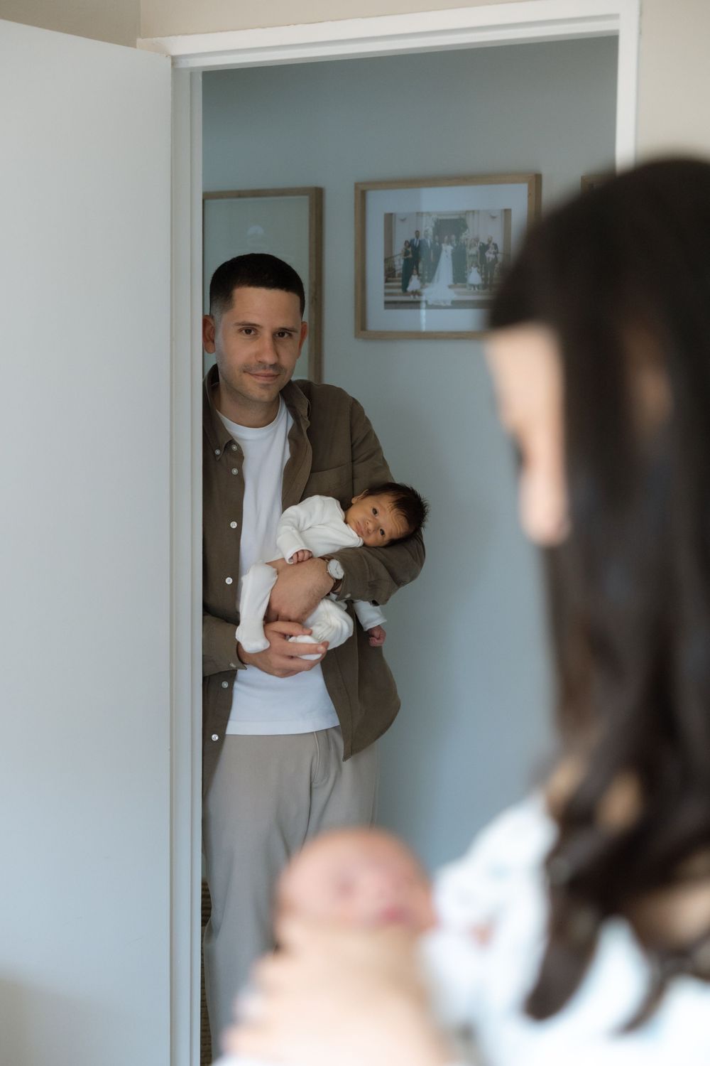 Father in olive shirt holds newborn baby in doorway while mother holds second twin in foreground.