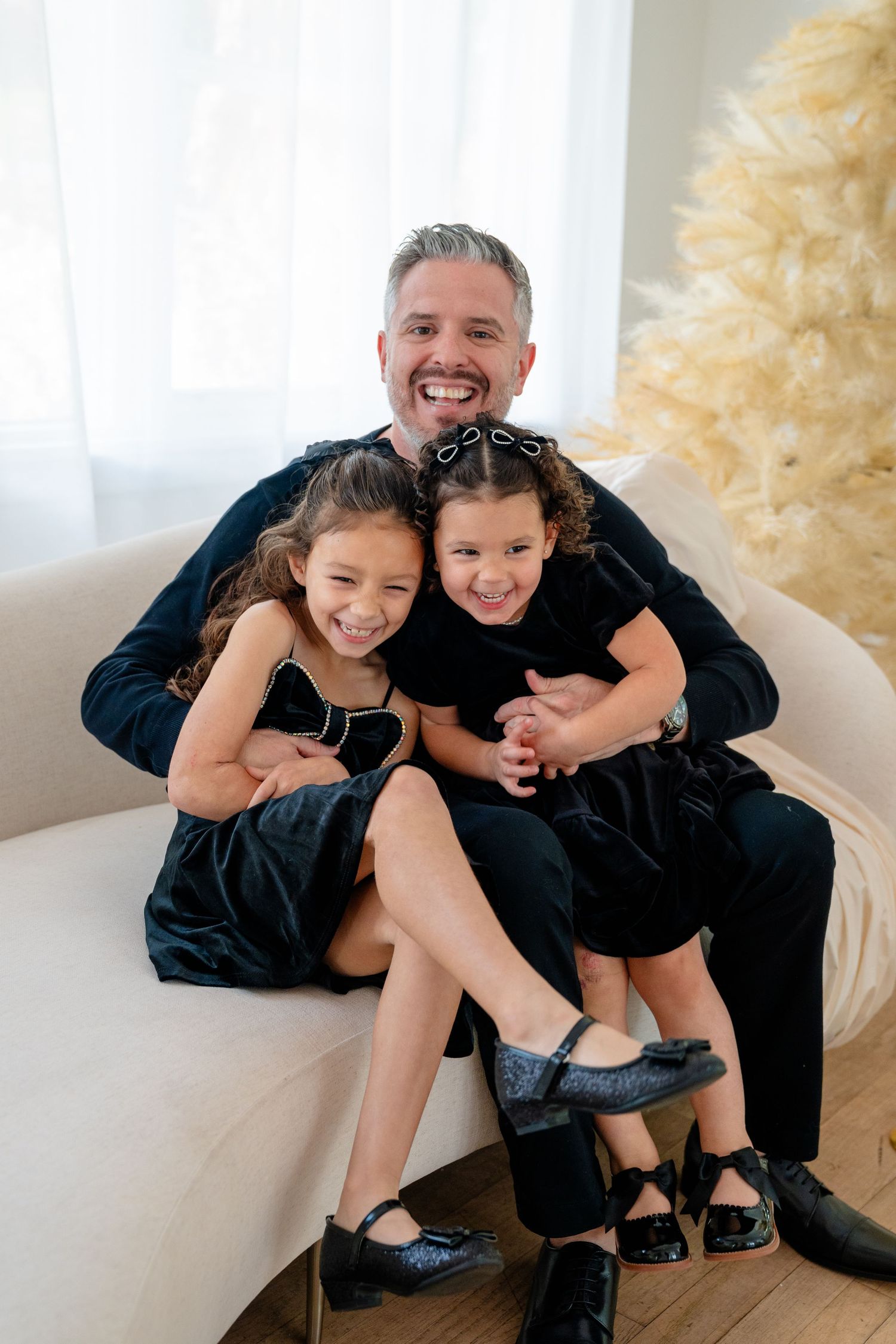 A smiling man in black sits on a sofa hugging two young girls in matching black dresses during a holiday photo.