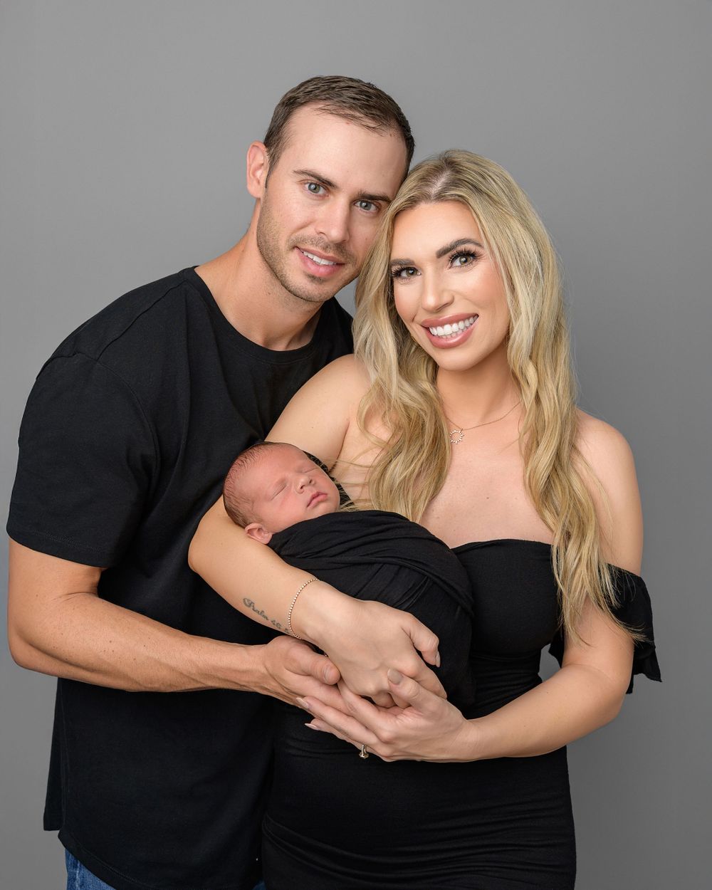 Couple dressed in black smiling together while holding their sleeping swaddled newborn in a studio photo.