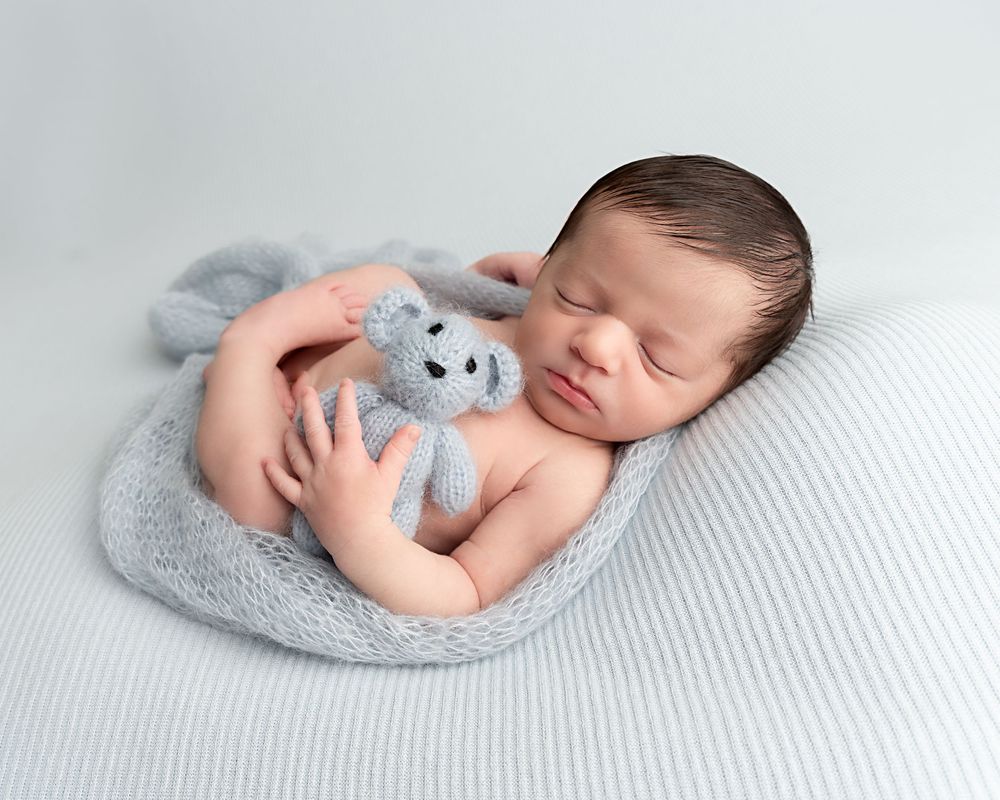 Peaceful sleeping newborn baby boy cuddling a grey knit teddy bear on a striped blanket.
