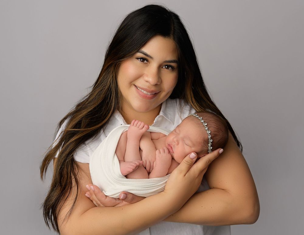 Young mother in white top tenderly cradling her sleeping swaddled newborn baby against her chest.