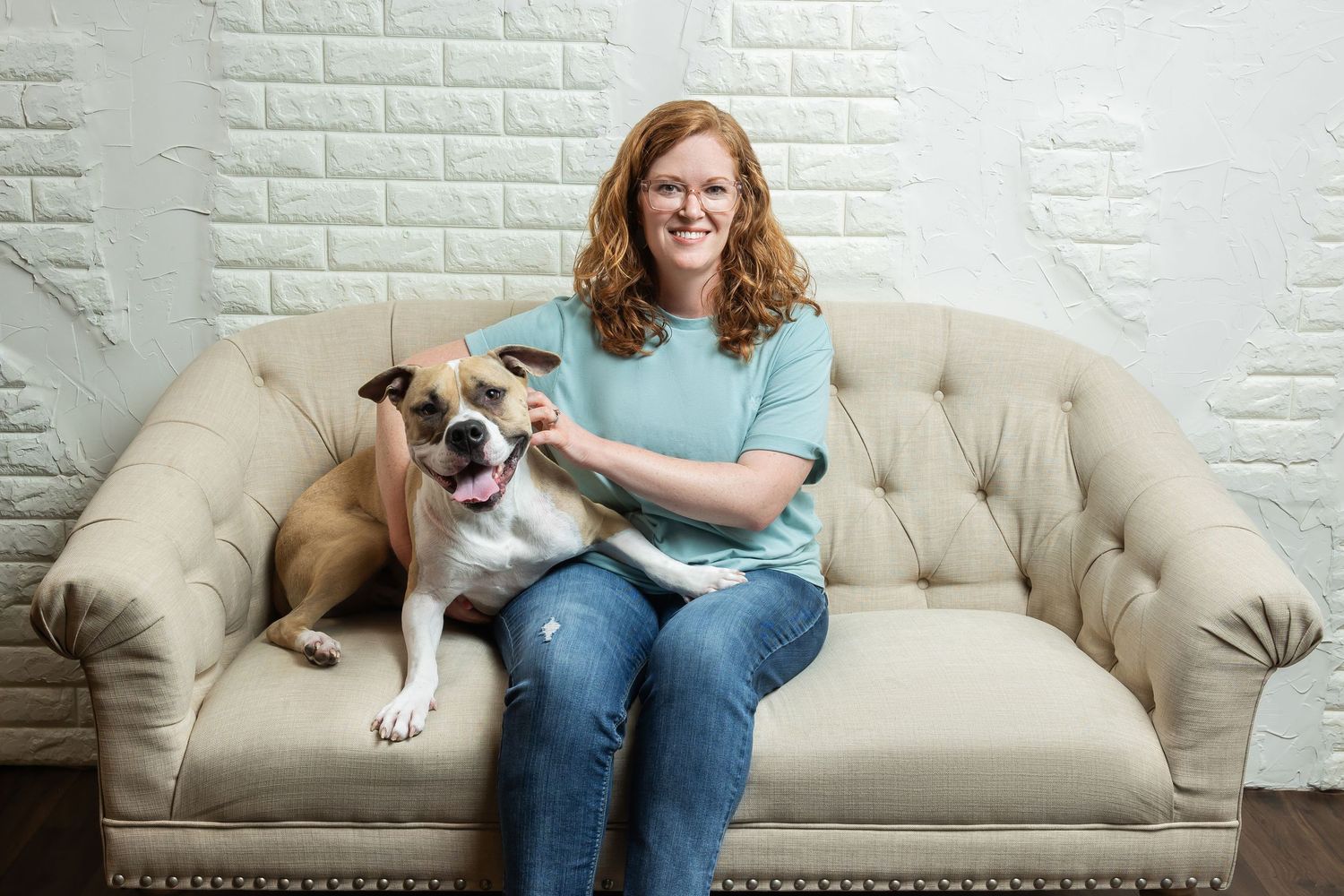 Woman with curly red hair smiling on a beige tufted sofa with a brown and white bulldog mix.
