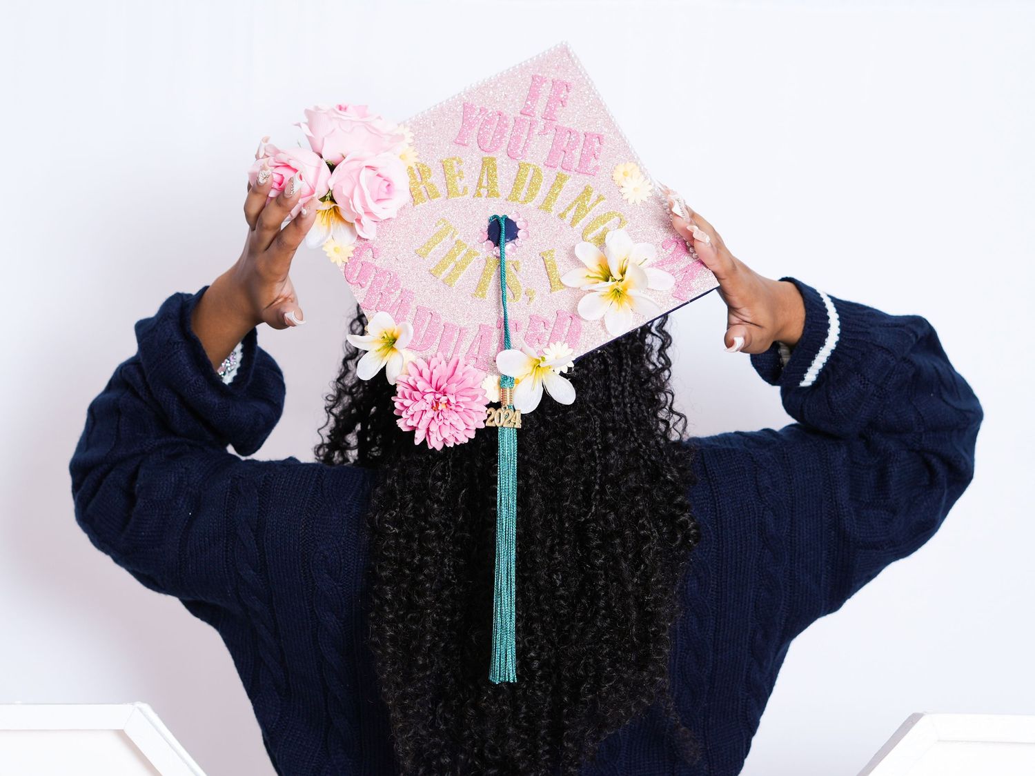 Graduate holding decorated floral graduation cap with gold text, wearing navy gown, with curly hair, against white background.