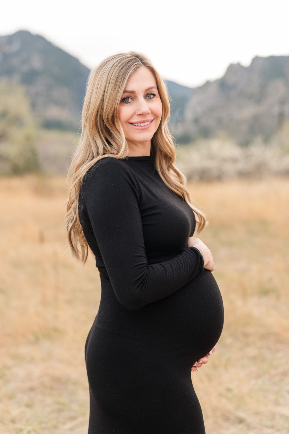 Smiling pregnant woman in black long-sleeve dress cradling baby bump in mountain field.
