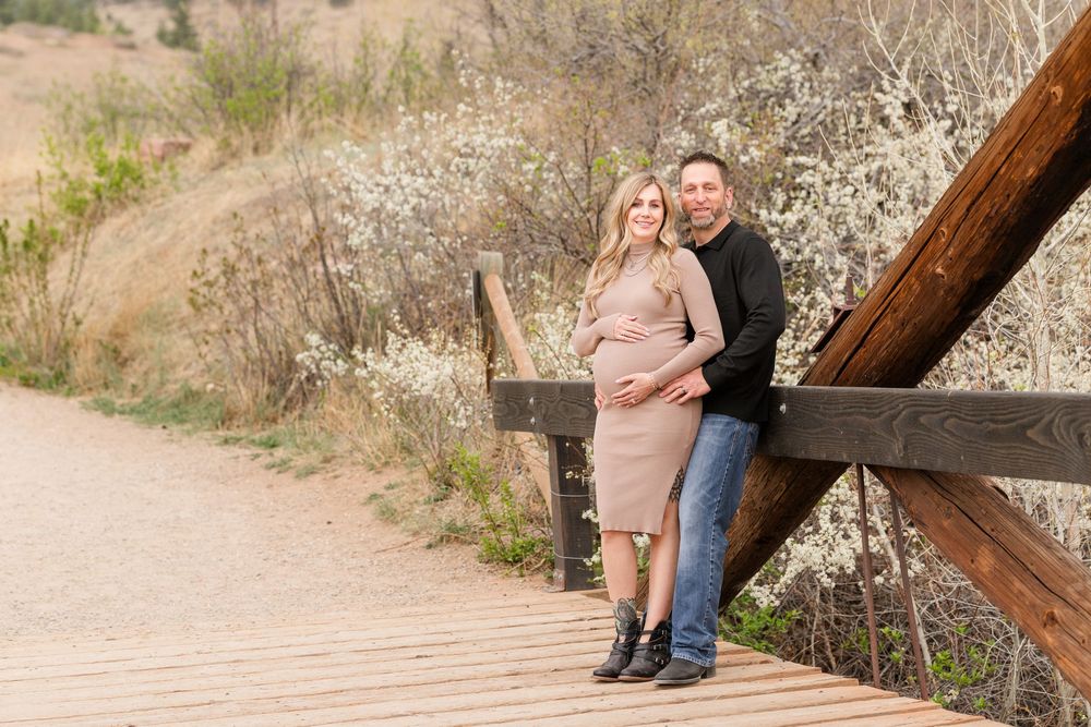 Expecting couple posing on wooden bridge surrounded by blooming shrubs, woman in blush dress, outdoor maternity portrait.