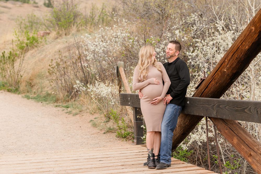 Pregnant couple embracing on a wooden bridge surrounded by blooming trees during an outdoor maternity photo session.