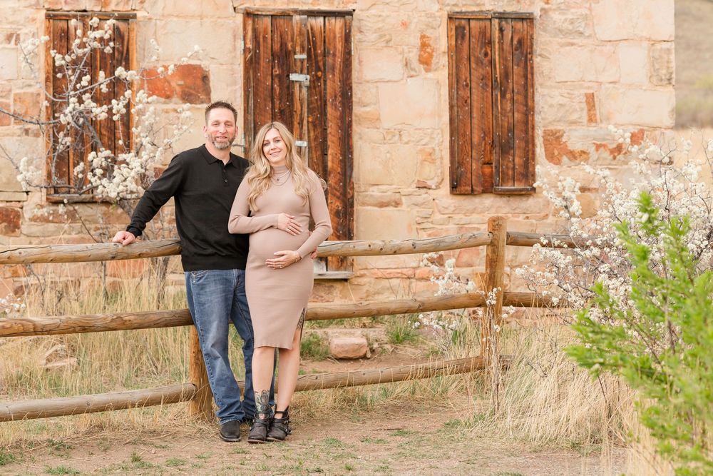 Smiling couple posing outdoors near rustic wooden fence and old stone building during maternity photo session.