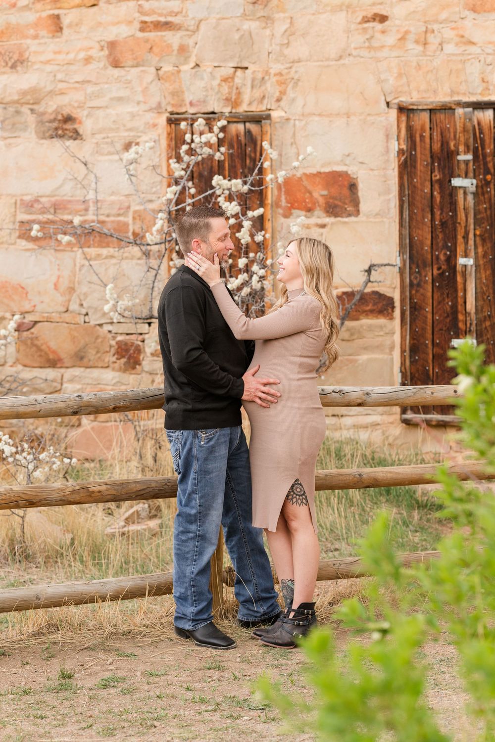 Couple embracing outdoors near rustic stone building and wooden fence, woman in beige dress, man in black sweater and jeans.