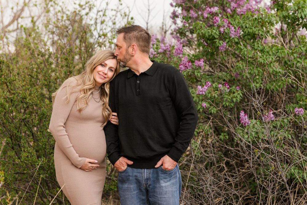 Pregnant couple embracing outdoors near blooming purple lilac bushes during a spring maternity photo session.