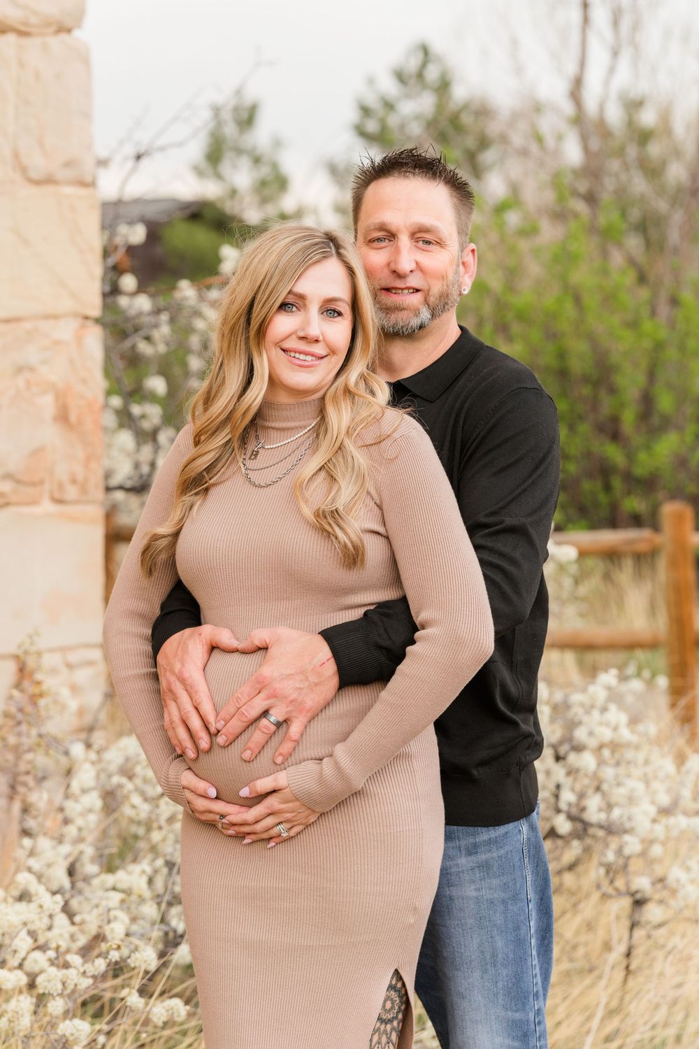 Smiling couple embraces outdoors, man in black shirt hugs pregnant woman in fitted beige dress.