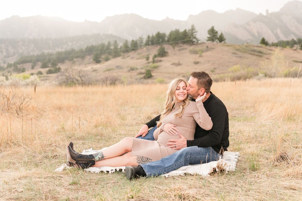 Couple sitting in golden field with mountains, man kissing pregnant woman's cheek during maternity photo session.