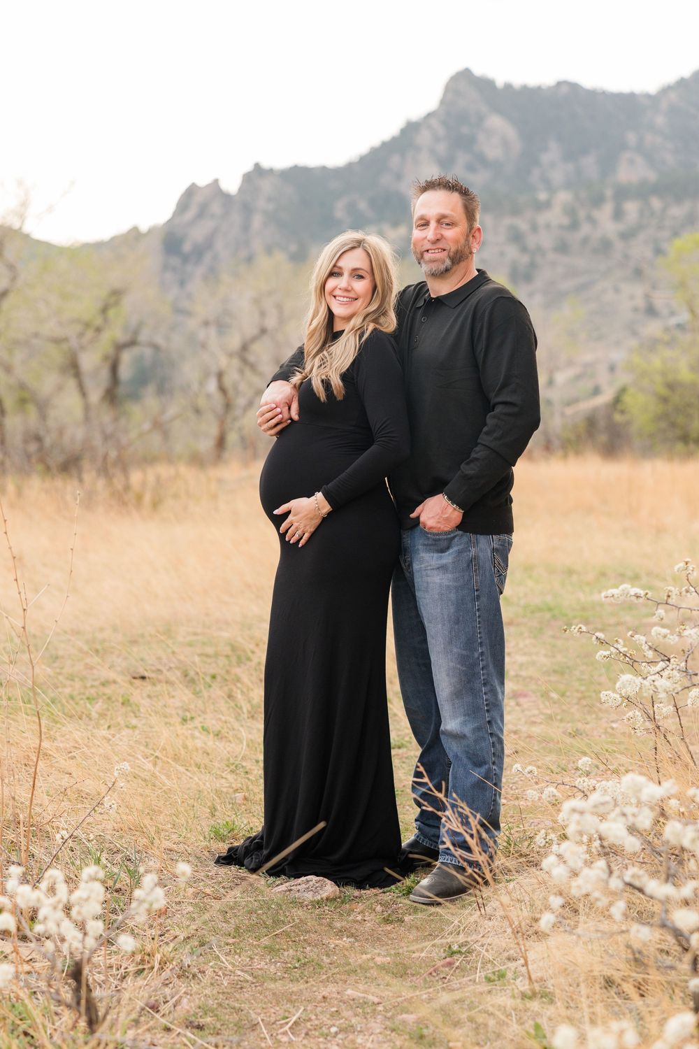 Pregnant couple posing outdoors in a mountain meadow, woman in black maternity gown, man in black sweater and jeans.