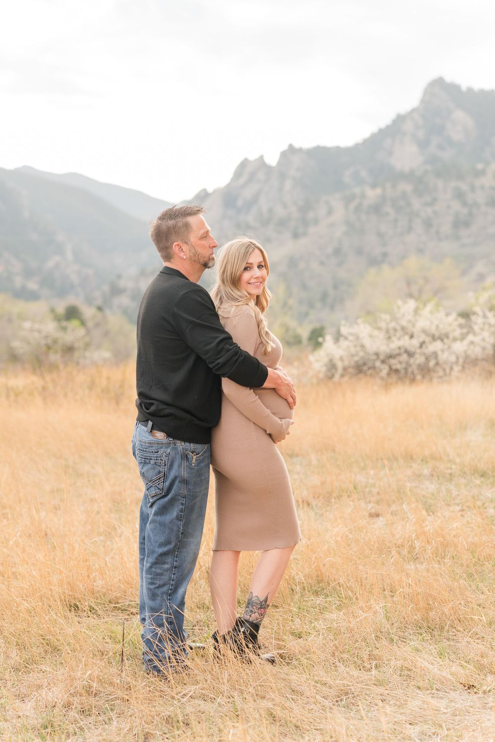 Couple embracing in golden field with rocky mountains backdrop, woman in beige dress smiling, maternity photo session.