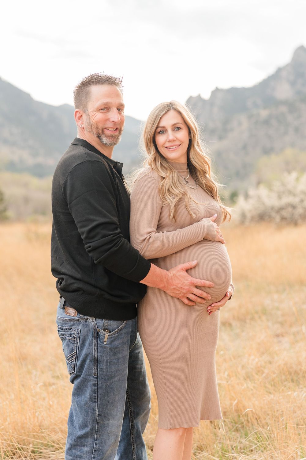 Couple posing in mountain field, pregnant woman in beige dress held by man in black sweater and jeans.