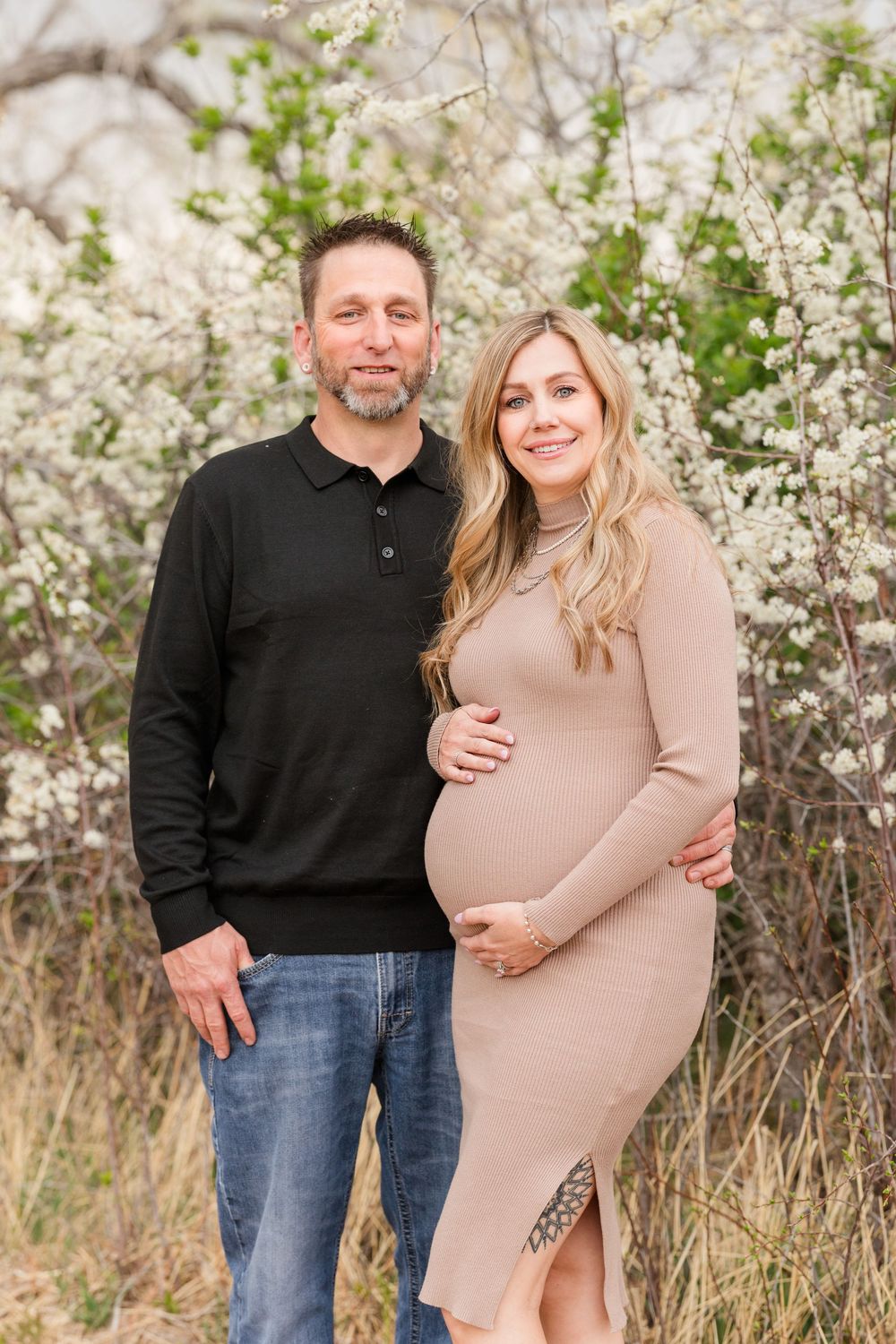 Couple poses outdoors among white blooming trees, woman in beige dress cradling pregnant belly.