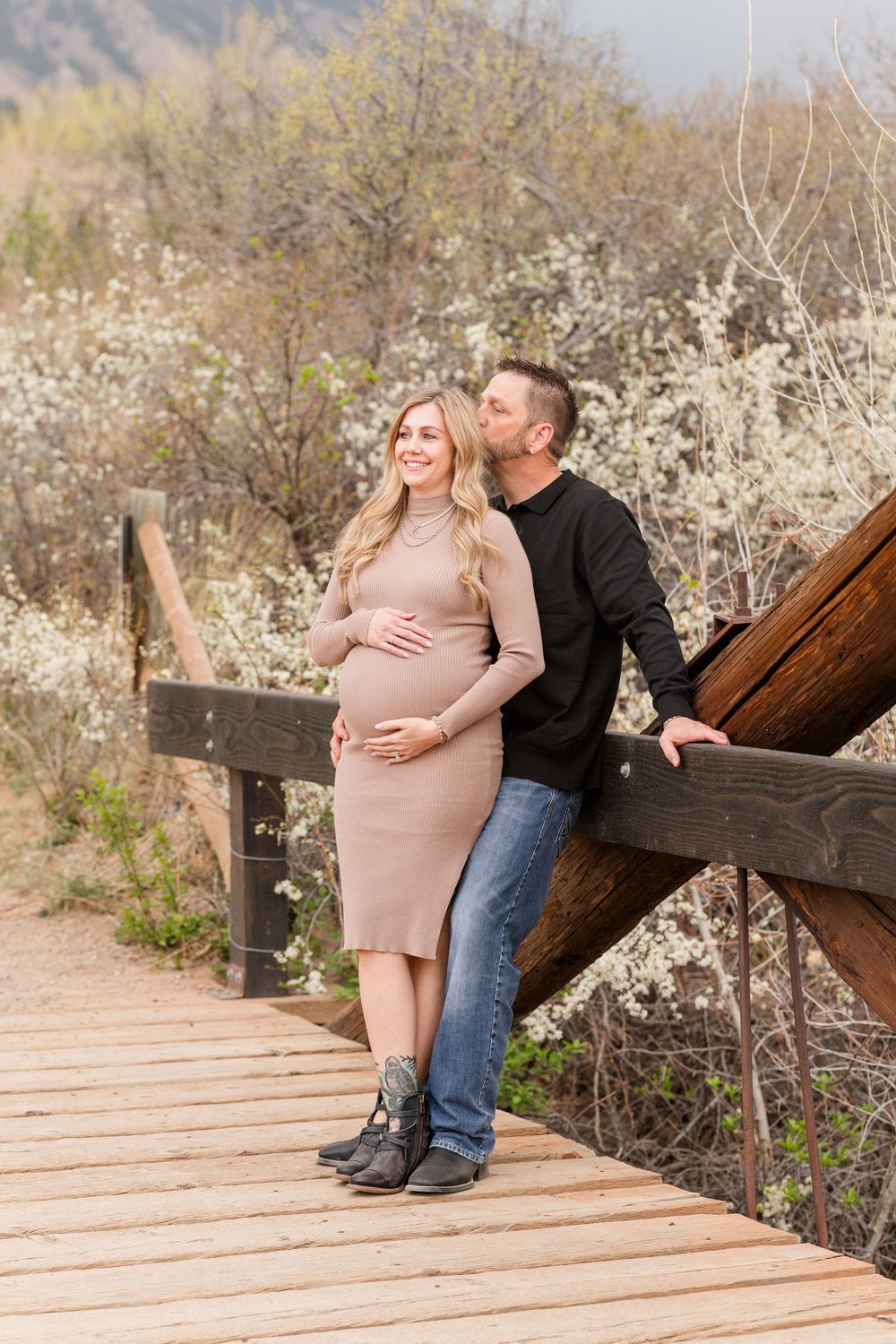 Pregnant couple on wooden bridge, man kissing woman's cheek as she cradles baby bump, surrounded by blooming spring trees.